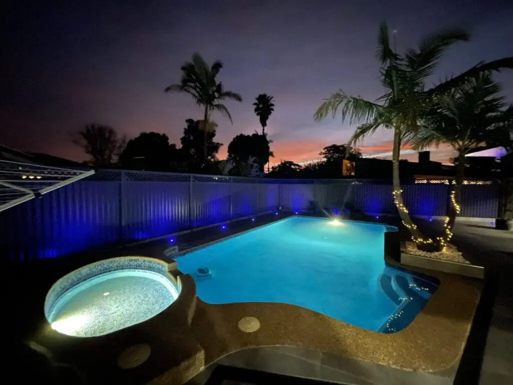 Outdoor pool and spa with blue lighting at dusk, framed by palm trees and a solar-powered home