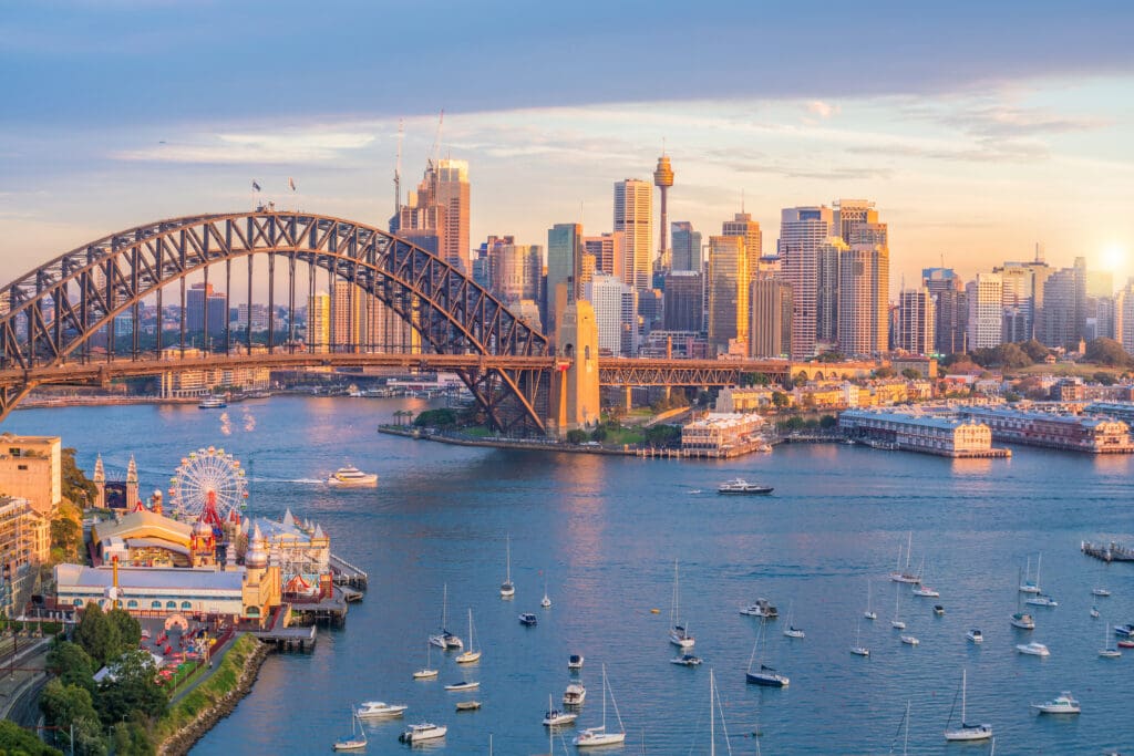 Aerial view of Sydney Harbour featuring the Sydney Harbour Bridge, city skyline, and Luna Park at sunset.