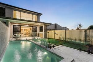 Modern backyard with a lit swimming pool, outdoor dining area, and a two-story house in the evening.